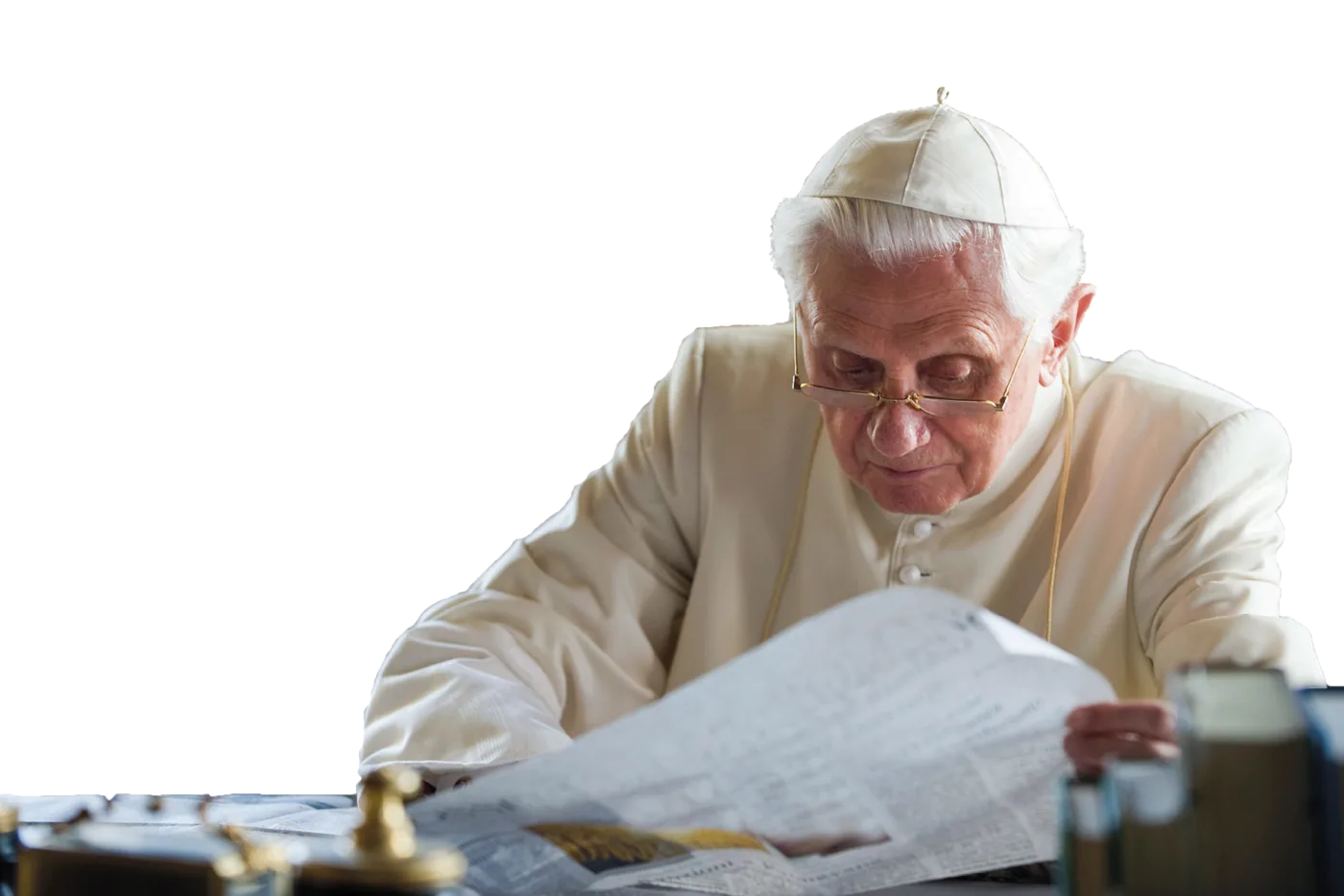 Papst Benedikt sitzt am Schreibtisch und liest Zeitung. Sein Blick ist dabei nach unten auf die Zeitung gerichtet.